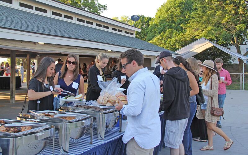 The image shows a group of people at an outdoor gathering, likely a picnic or barbecue. There's a food serving area with various dishes. People are seen serving themselves. The atmosphere seems relaxed and social, with a focus on food and interaction.